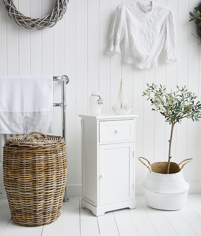 Maine white bathroom cabinet. White Furniture for storage with a cupboard and drawer. Photograph shows the cabine in a white bedroom beside the Casco Bay laundry basket