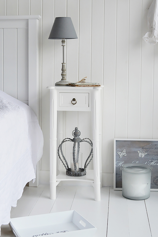 A white bedside table with a single drawer with an antique brass drop pull handle and a bottom shelf.

With its simplicity and elegance it will suit well in any home. The tall narrow table features rounded corners, engraved spindle legs and a bottom shelf to set books or a basket.