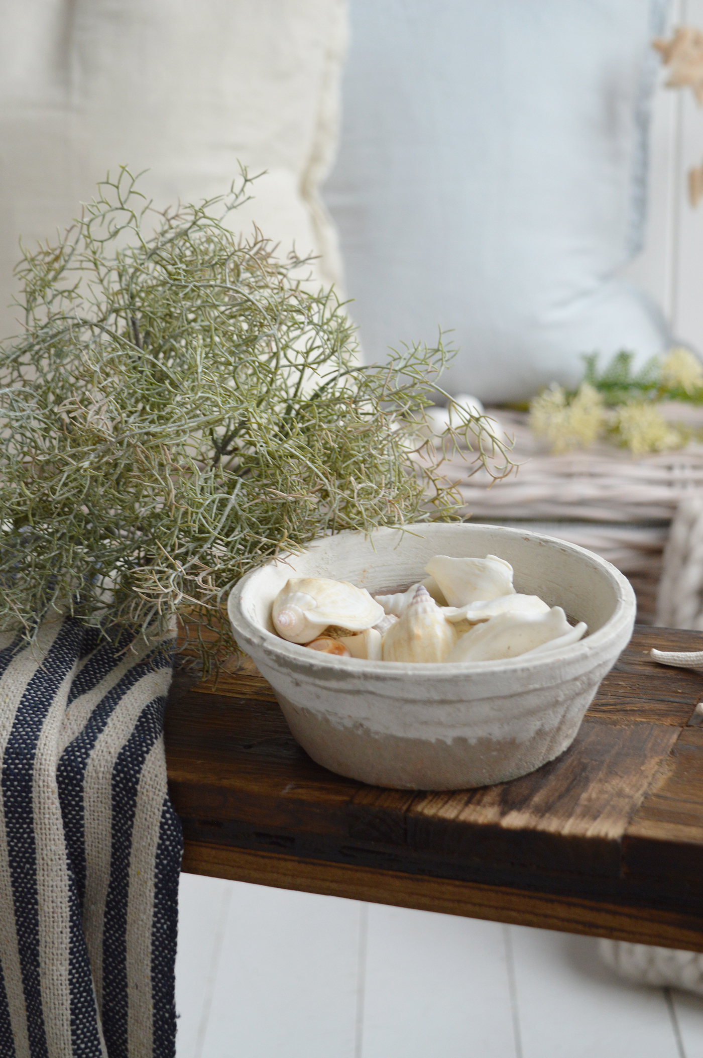 Ceramic bowl filled with seashells styled with coastal foliage and striped linen on rustic bench in Hamptons beach house interior