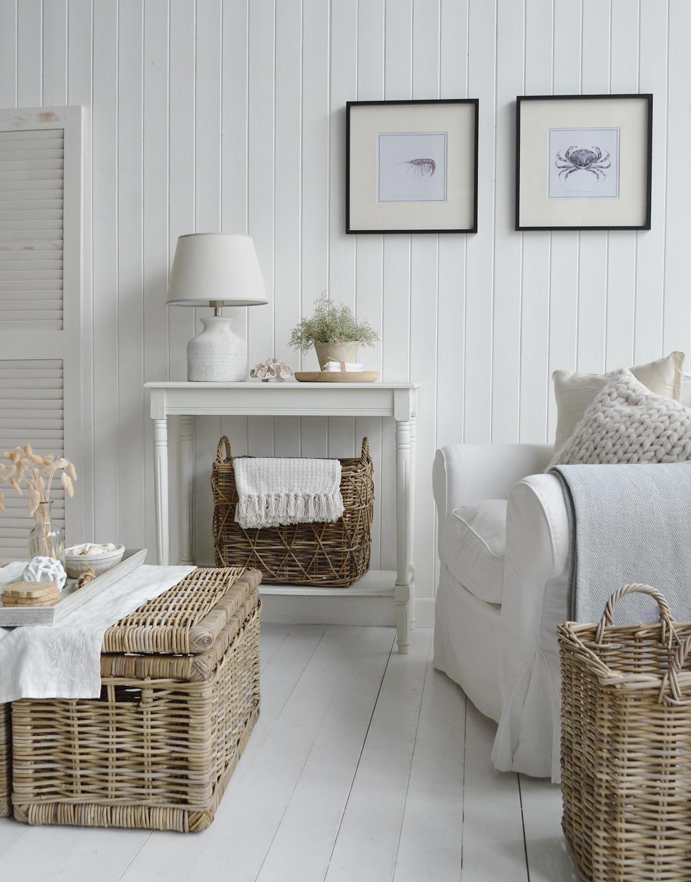 This image shows a bright, coastal-style living space with white shiplap walls and floorboards. A wicker coffee table with a linen runner sits in front of a white armchair, which is draped with a light gray throw and chunky knit pillow. Behind it, a small white console holds a wicker basket and simple decor, while two framed sea creature prints hang on the wall above. Natural textures and soft neutrals give the room a calm, beachy feel.