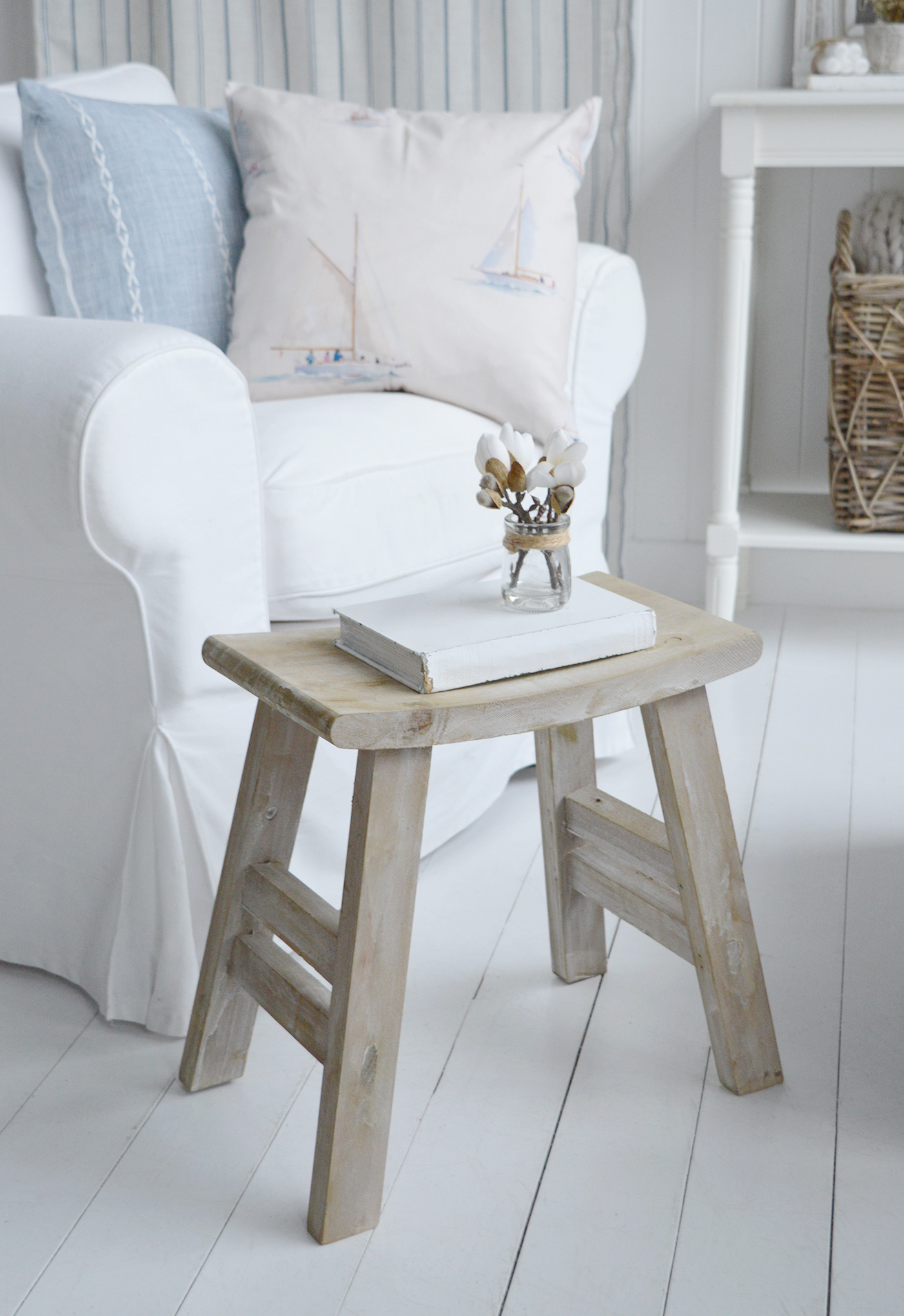 A light-washed Pawtucket wooden stool styled beside a white linen armchair with soft blue nautical cushions, bringing relaxed coastal charm and effortless Hamptons style to bright, neutral living room interiors.