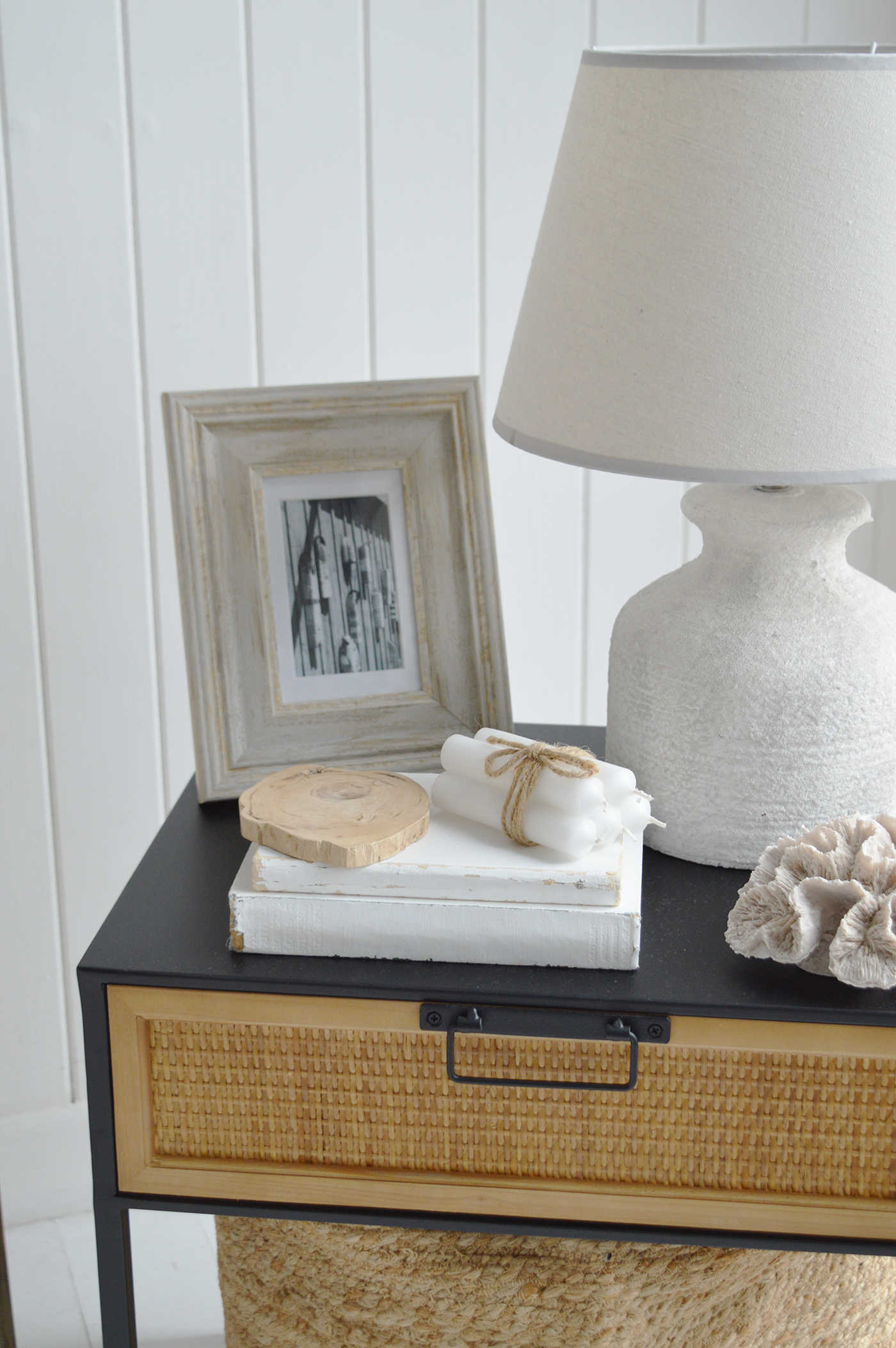 Next to the sofa is a small black-framed side table with woven rattan drawer detailing.Decor items on the table: A ceramic lamp with a fabric shade, a picture frame with a monochrome photo, a small stack of books, a decorative shell, and a piece of coral all contribute to the coastal theme.