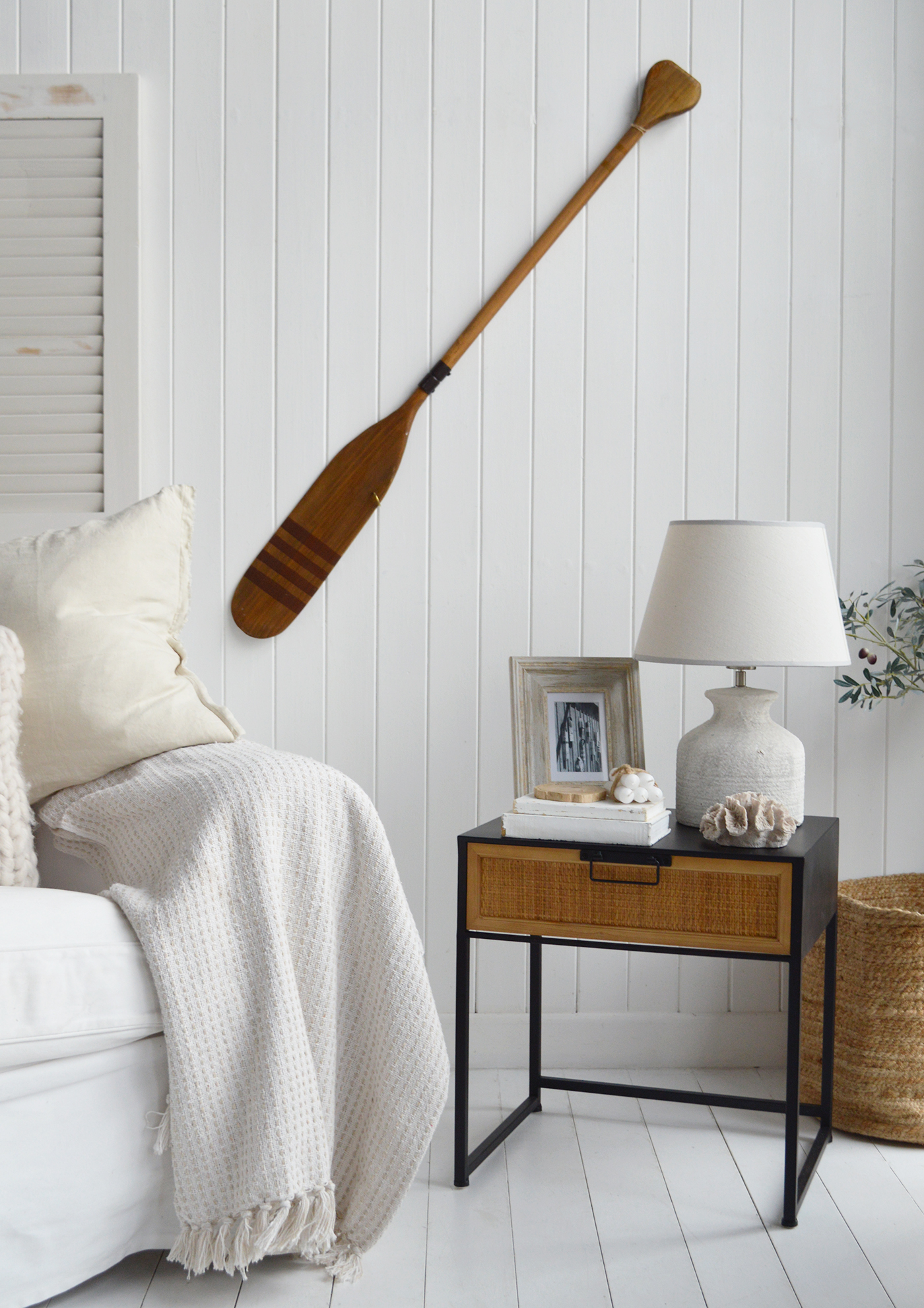 This photo captures a serene coastal-inspired corner, styled with soft neutrals and natural textures. A wooden oar mounted on the white paneled wall adds a nautical touch, while the rattan-drawer side table holds a rustic lamp, framed photo, and seashell accents. Cosy textiles, like the tasseled throw and cushion, create a relaxed, inviting atmosphere, perfectly balanced by the woven basket nearby.