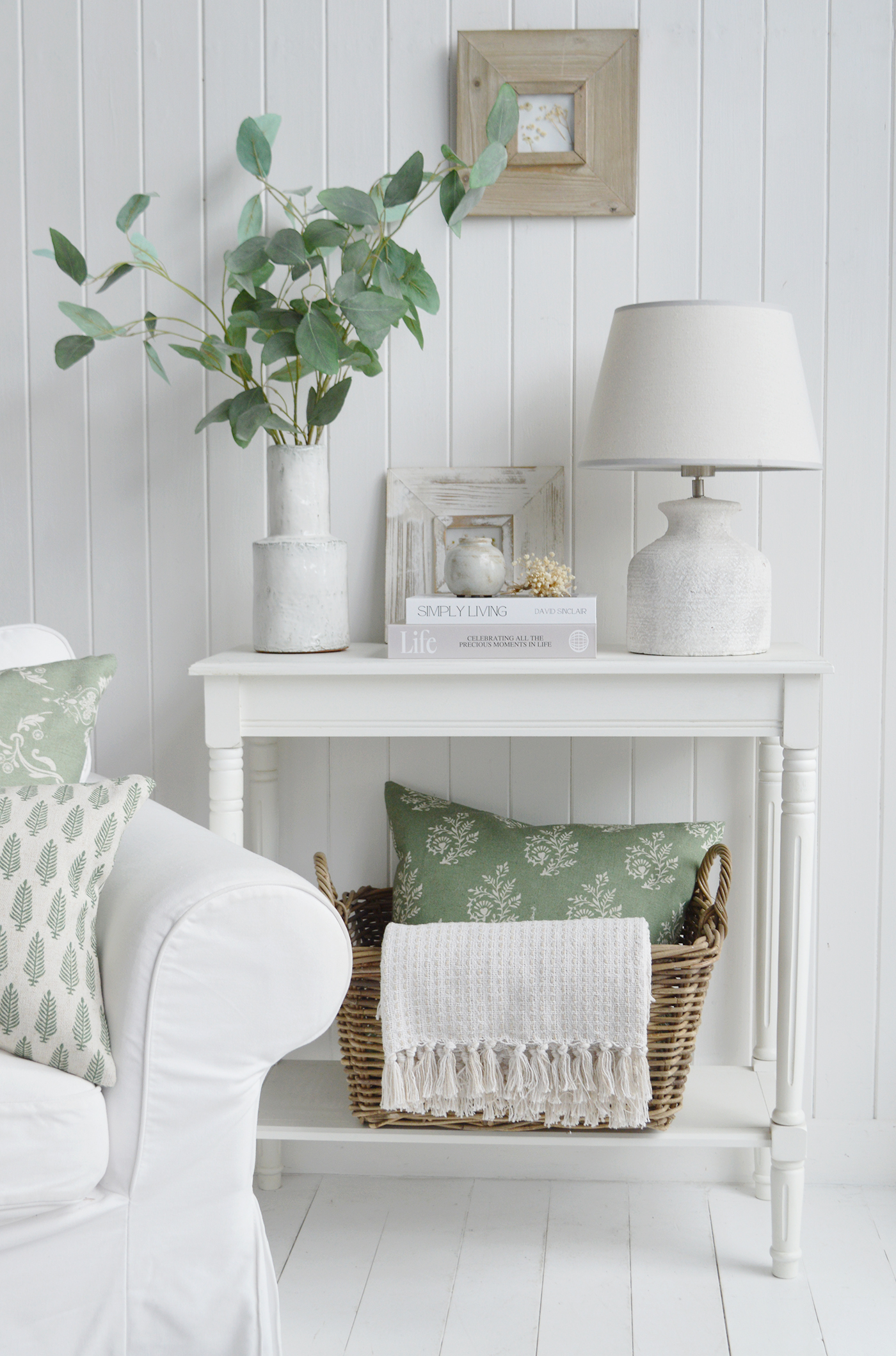A carefully curated blend of soft sage greens, natural textures and timeless white furnishings creates a beautifully balanced coastal interior. The crisp white console table provides the perfect backdrop for layered styling, with textured ceramics, botanical stems and weathered wooden accents adding warmth and character. A woven basket filled with a cosy throw and patterned cushion introduces depth and comfort, while the fresh green tones bring a subtle connection to nature. Inspired by relaxed New England living, this elegant arrangement demonstrates how simple accessories, natural materials and a restrained colour palette can transform a space into a calm and inviting retreat.