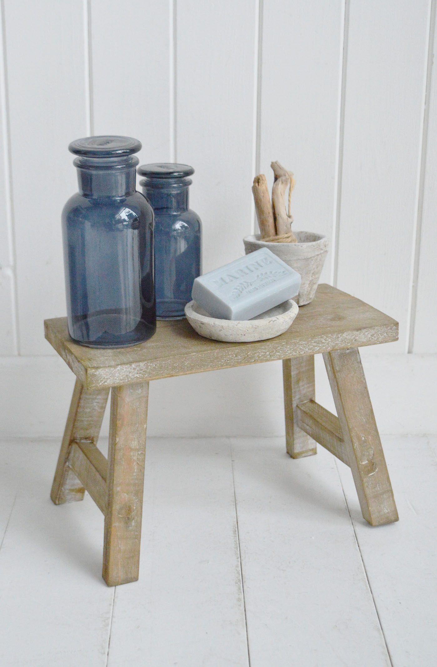 The Pawtucket small wooden stool in a softly weathered finish is perfect for styling in coastal New England interiors, beautifully displaying blue glass bottles and natural stone accessories for relaxed beach house décor.
