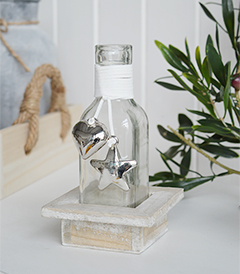 A decorative square bottle with a hanging silver mirrored chunky heart and star in a driftwood style wooden crate for New England coastal and country interiors and white furniture