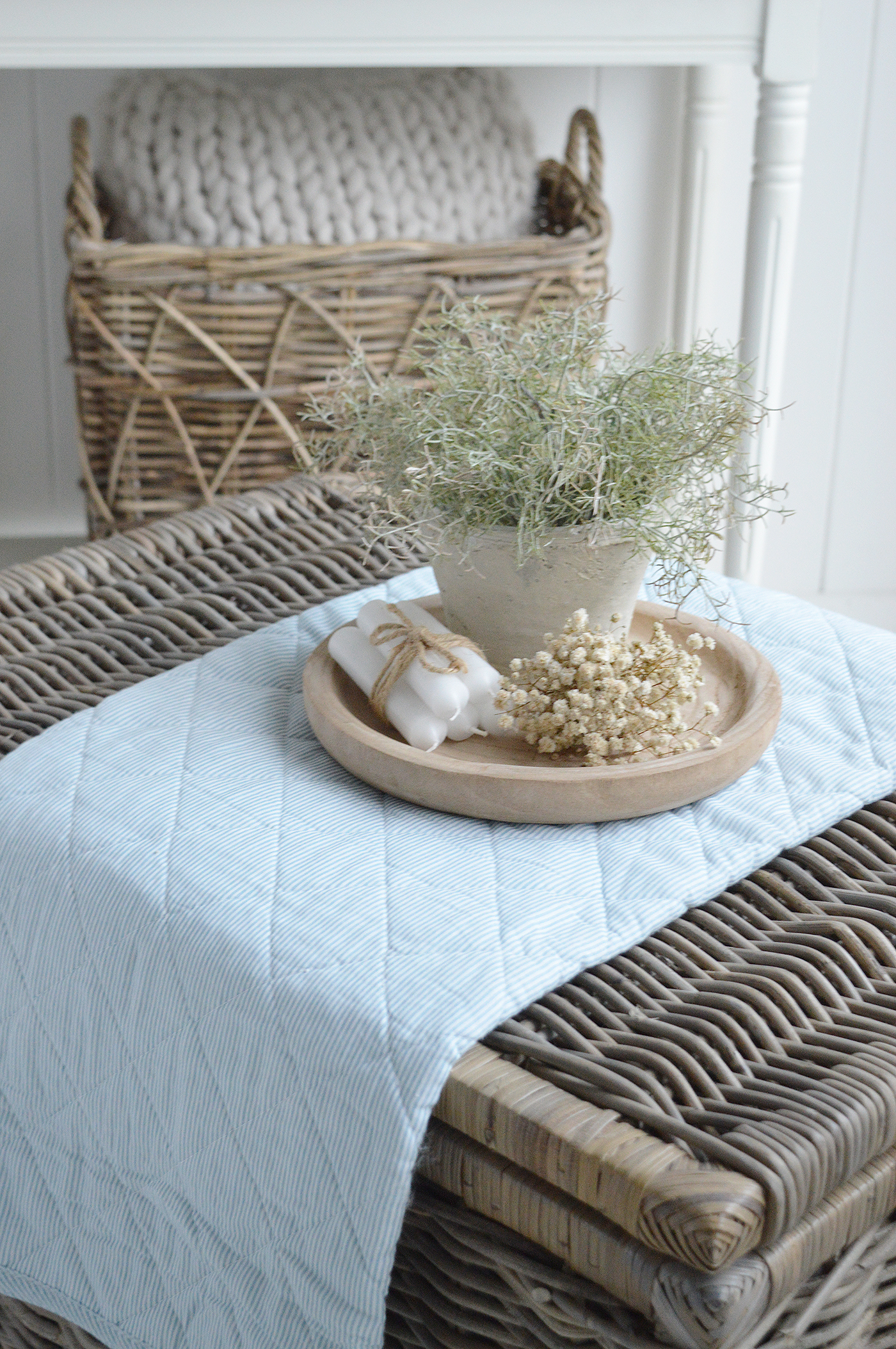 A softly styled coastal vignette featuring a woven rattan trunk, Piermont pale blue striped quilted throw, natural wooden tray, and neutral ceramics, creating a relaxed Hamptons-inspired look perfect for light, airy coastal and modern country interiors.