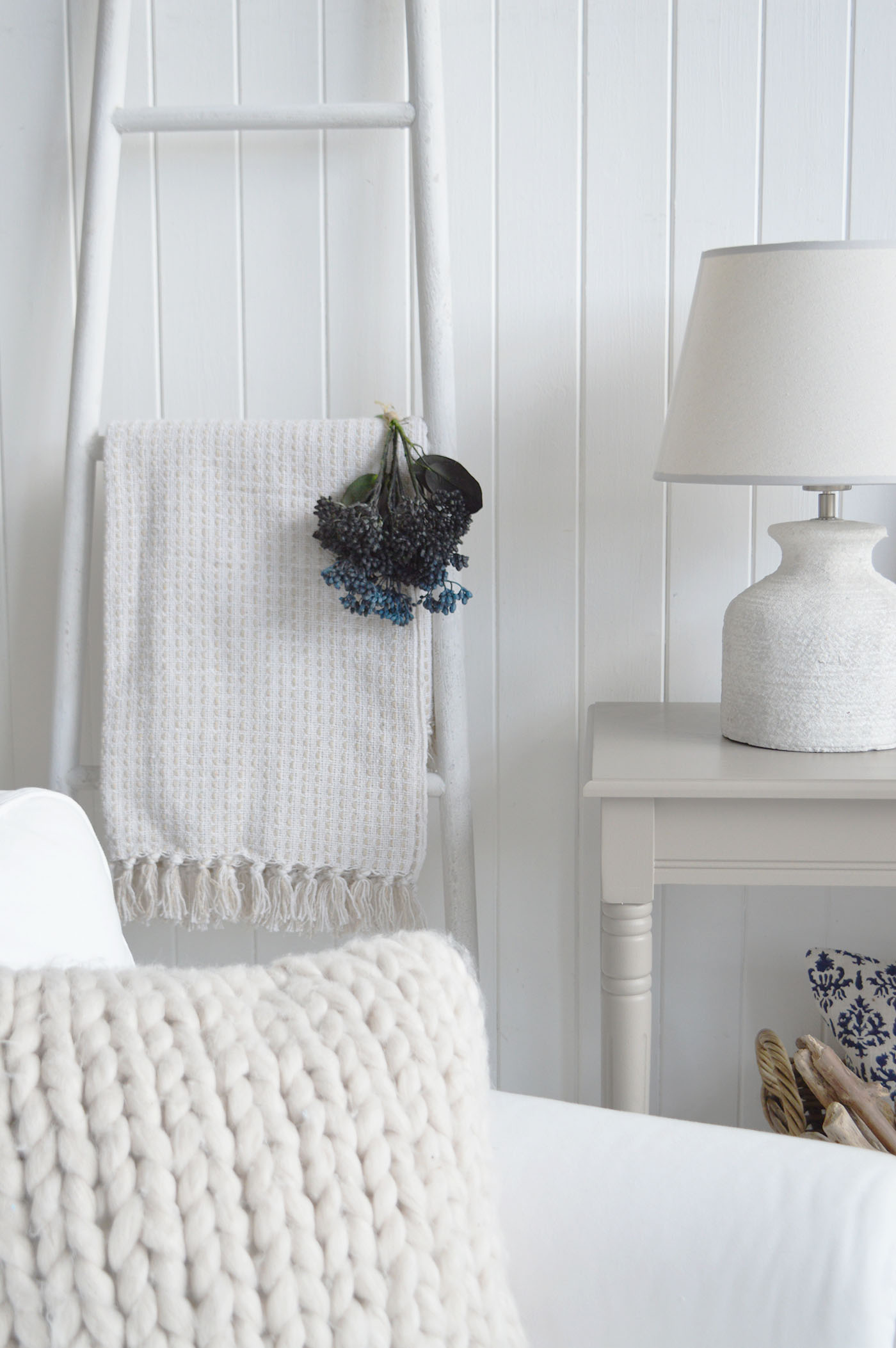 A serene coastal beach house interior featuring white shiplap walls, a textured ceramic table lamp, soft neutral throws, and a rustic ladder display, creating a relaxed New England coastal style perfect for calm, light-filled living spaces.