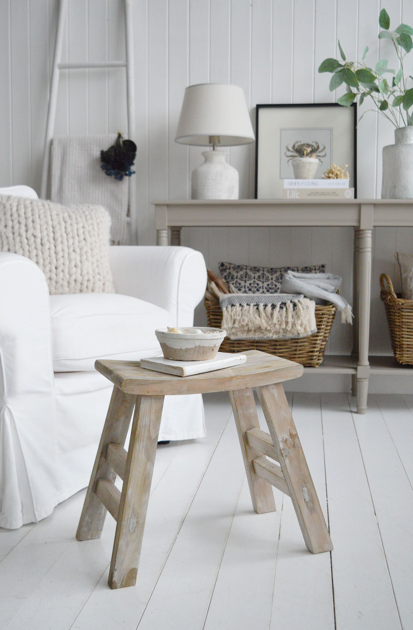 This Pawtucket rustic wooden stool with a softly weathered finish adds natural warmth and texture to neutral coastal interiors, beautifully styled beside a white armchair and shiplap walls for relaxed New England beach house living.