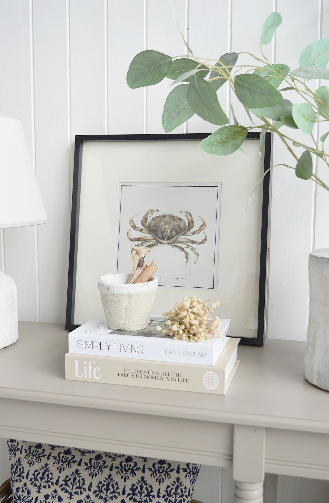 A coastal console table styling with neutral books, textured pottery, driftwood accents and a framed crab illustration, set against white shiplap walls to create a calm New England beach house interior with natural textures and relaxed coastal charm.