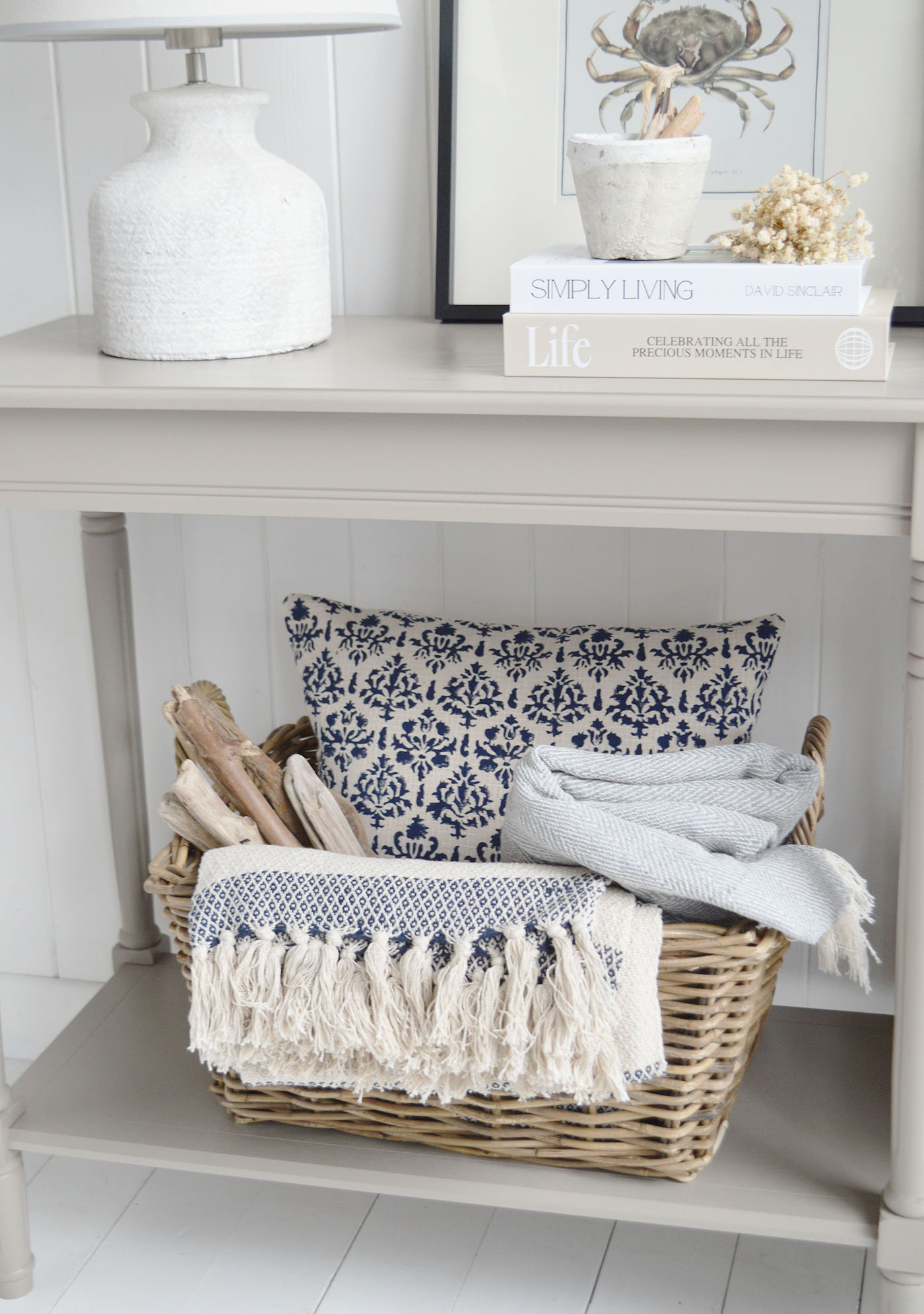 A beautifully styled coastal beach house console table featuring a textured ceramic lamp, woven basket with neutral throws, driftwood accents and blue patterned cushions, creating a relaxed New England coastal interior with layered natural textures and soft neutral tones.