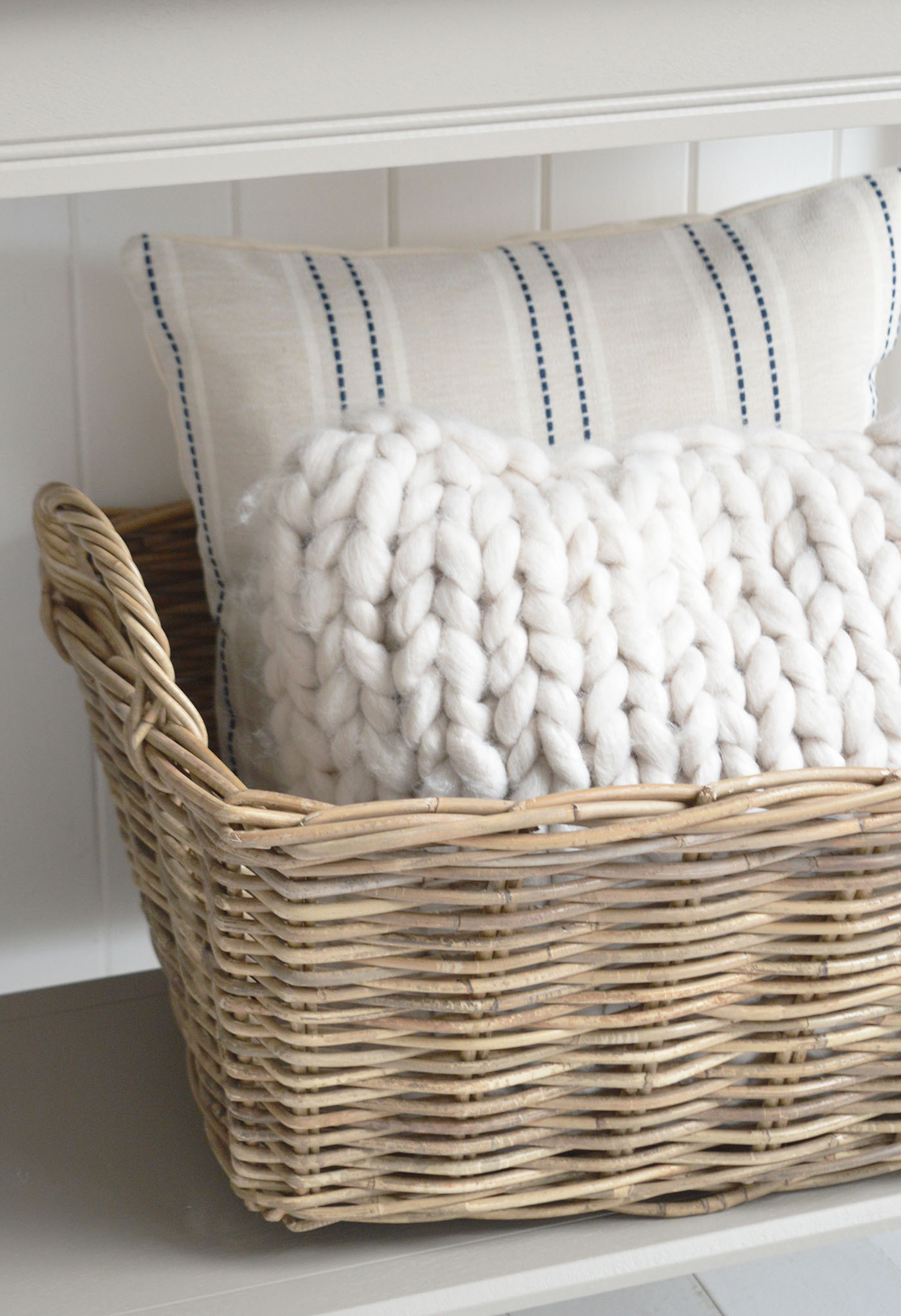 A woven wicker basket filled with soft neutral throws and a blue stripe coastal cushion, perfectly styled against white shiplap walls to create a relaxed New England beach house interior with natural textures and calm coastal tones.