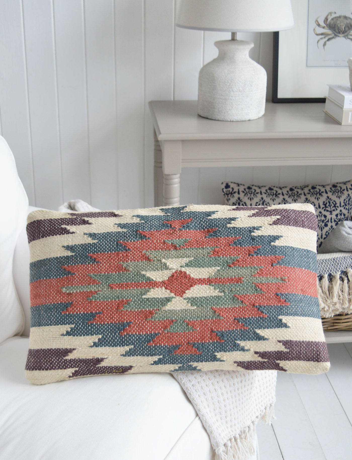 A textured kilim style cushion in muted coastal tones of rust, blue and sage styled on a soft neutral sofa, bringing relaxed beach house charm and layered coastal interior texture to this light-filled New England inspired living space.