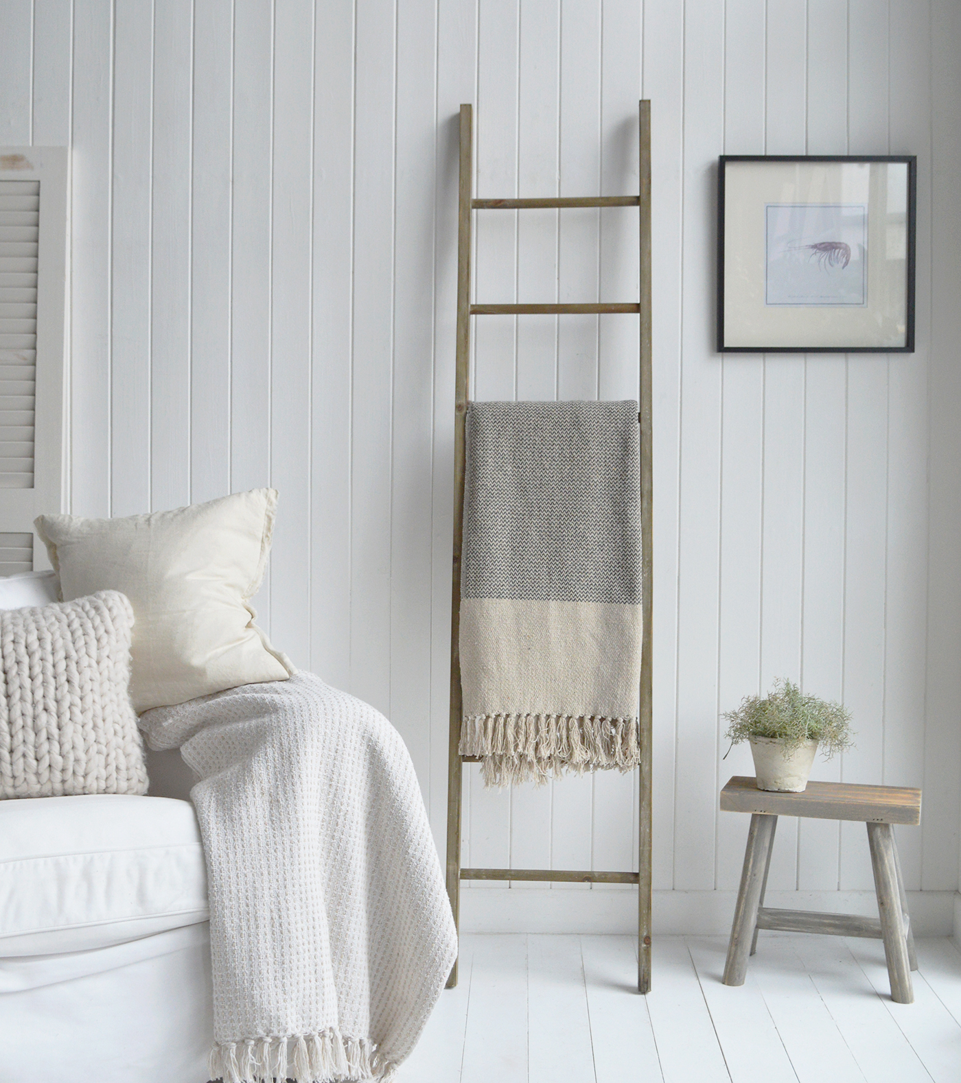 This space blends Hamptons elegance with New England coastal charm and farmhouse warmth. White panelled walls and pale tones create a light, airy feel, while natural textures—like the knit cushion, fringed blanket, and rustic ladder—add depth and comfort. Simple accents, such as the wooden stool with greenery and subtle artwork, complete the serene and timeless look.