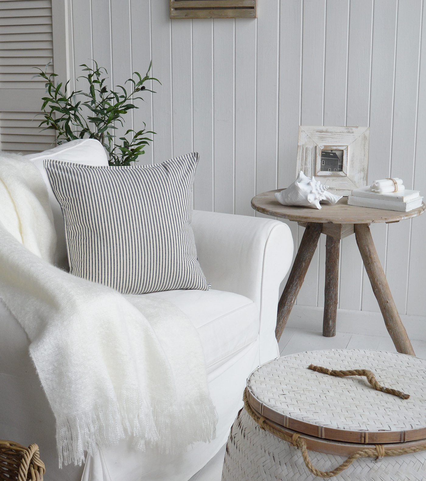 Navy pinstripe rhode island cushion cover for New England beach house and coastal interiors. Photograph shows a living room with a white sofa, the driftwood table, Nantucket white basket table and coastal interio decor