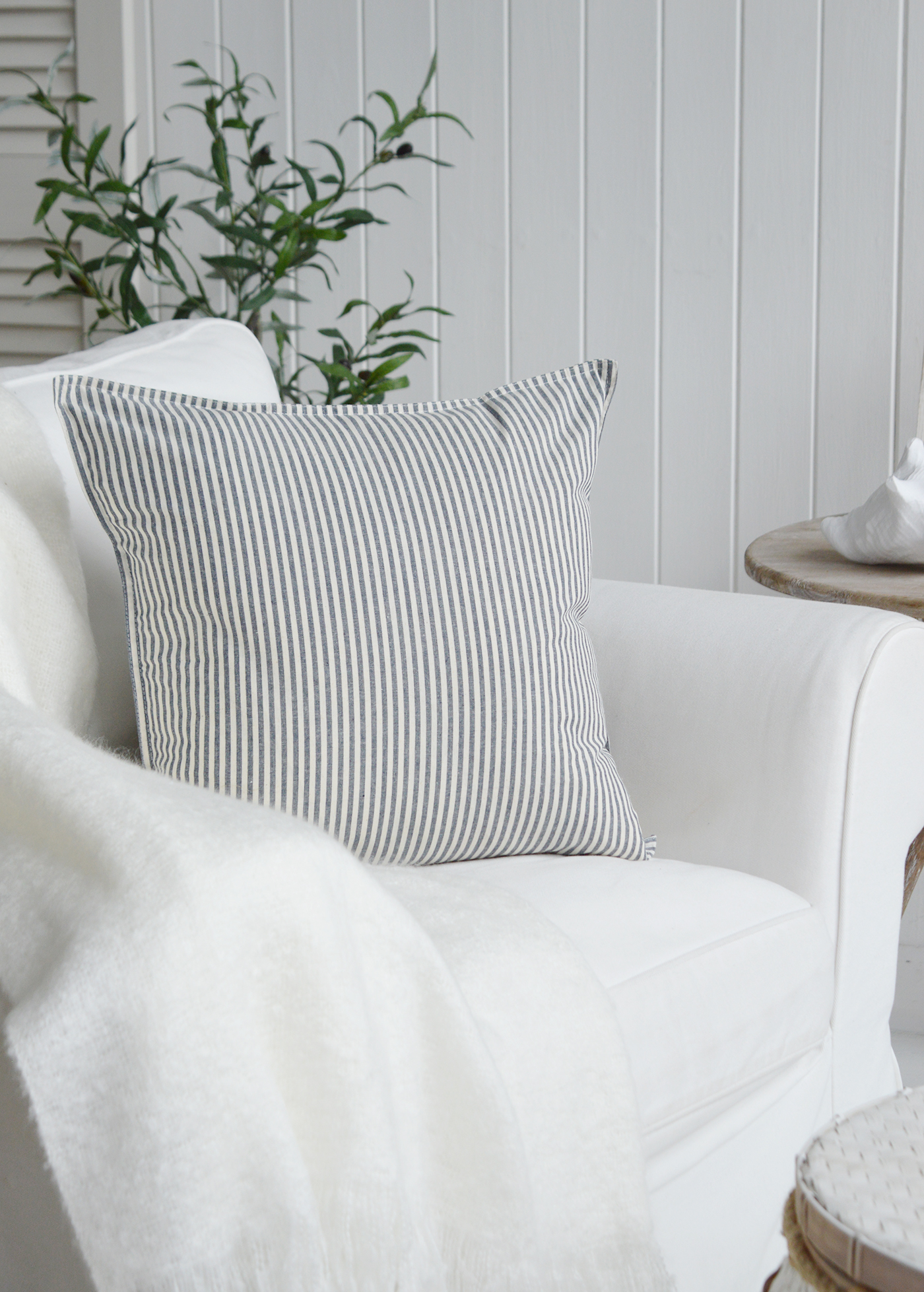 Navy pinstripe rhode island cushion cover for New England beach house and coastal interiors. Photograph shows a living room with a white sofa, the driftwood table, Nantucket white basket table and coastal interio decor