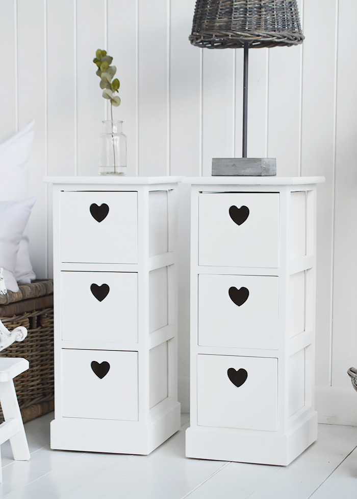 A pair of narrow white bedside tables with three drawers and cut out hearts 25cm wide