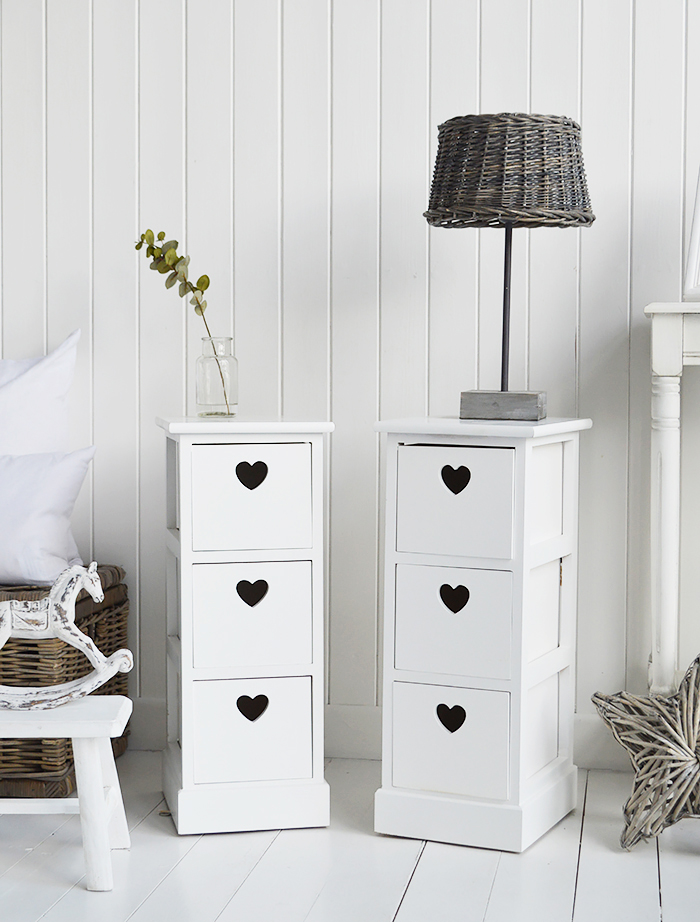 A pair of narrow white bedside tables with three drawers and cut out hearts 25cm wide