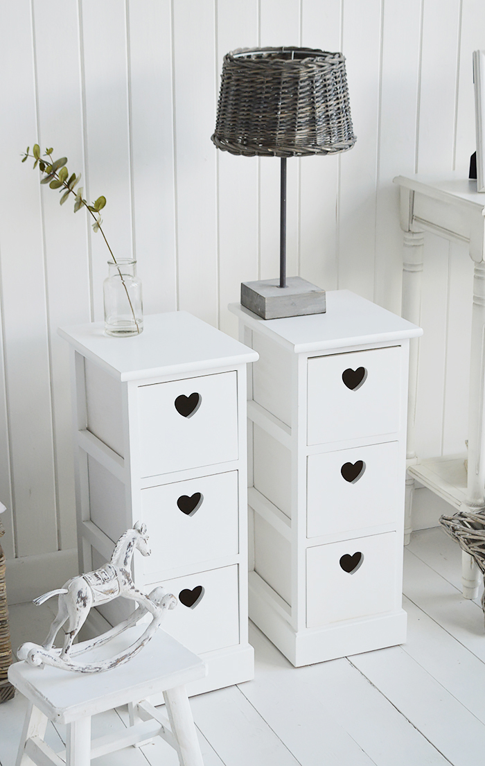 A pair of narrow white bedside tables with three drawers and cut out hearts 25cm wide