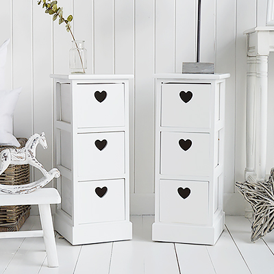 A pair of narrow white bedside tables with three drawers and cut out hearts 25cm wide